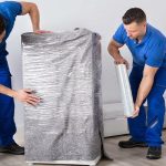 Two workers in blue uniforms securely wrap a large item in plastic for transport. Safe Packaging in Shipping and Logistics.